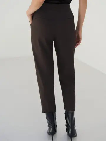 A person shown from the back wearing dark brown high-waisted ankle-length trousers and black pointed-toe heeled boots against a light gray studio background.