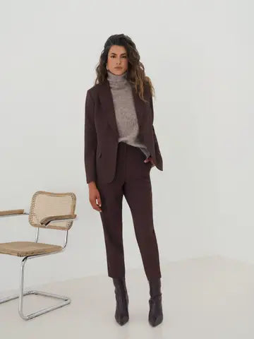 A woman models a dark brown tailored suit consisting of a blazer and slim trousers, styled with a light gray turtleneck sweater and dark brown pointed-toe ankle boots. She stands in a minimalist studio setting with a modern chair on the left.