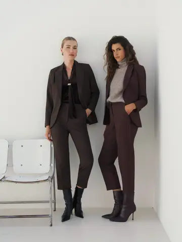 Two women modeling dark tailored suits in a minimalist white studio setting. One wears a black-and-brown blazer with matching trousers and black ankle boots, and the other wears a dark brown blazer with matching trousers over a light gray turtleneck and black ankle boots.