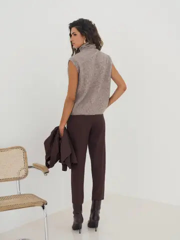 A woman stands in a studio wearing a sleeveless taupe-gray textured mock-neck top, dark brown trousers, and black ankle boots, holding a dark brown jacket. A woven chair is partially visible on the left.
