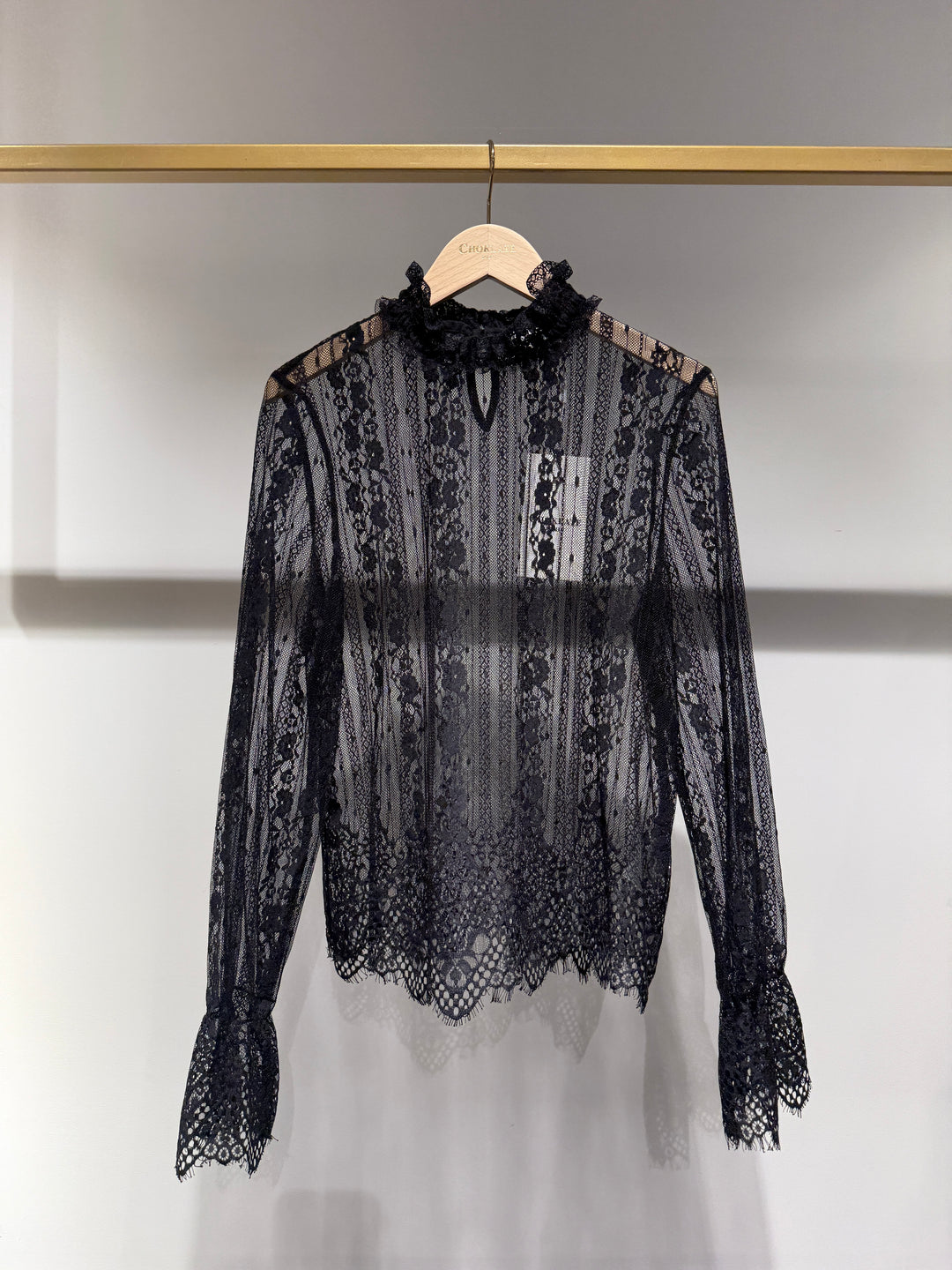 A black sheer lace long-sleeve blouse with a ruffled mock neck and scalloped hem, hanging on a wooden hanger against a white wall.
