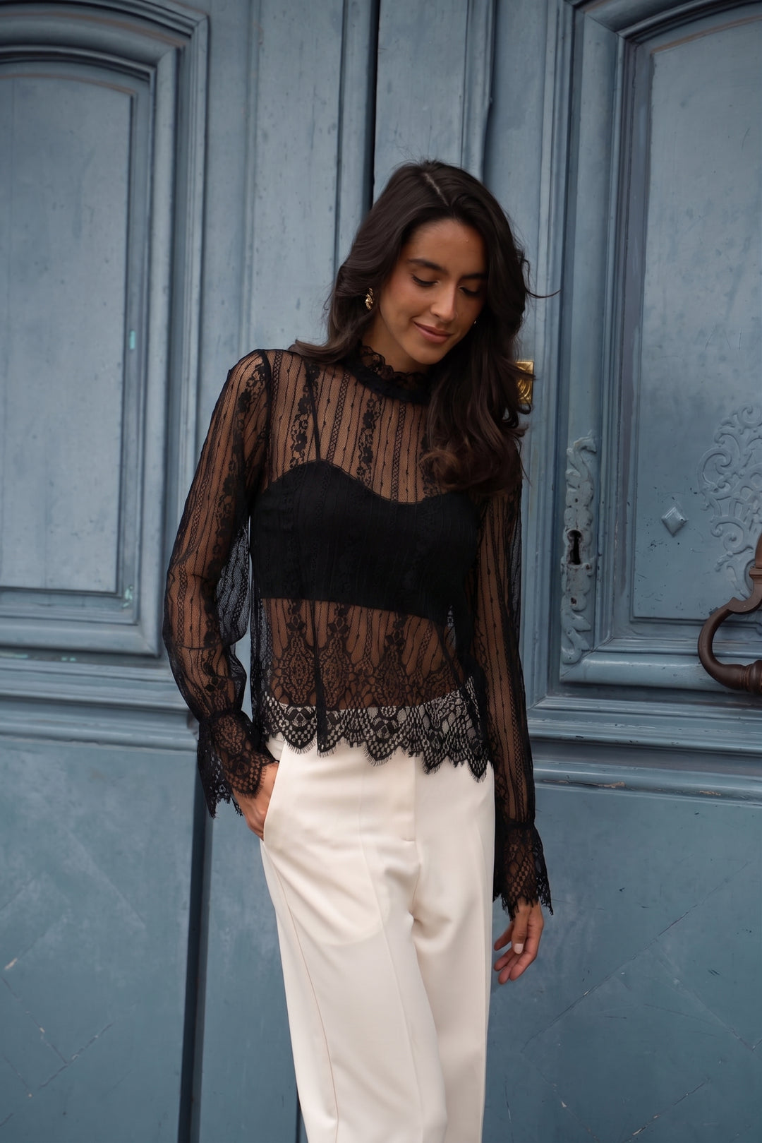 A woman wearing a sheer black lace long-sleeve top with a solid black sweetheart bodice, paired with off-white trousers, standing in front of a blue wooden door.
