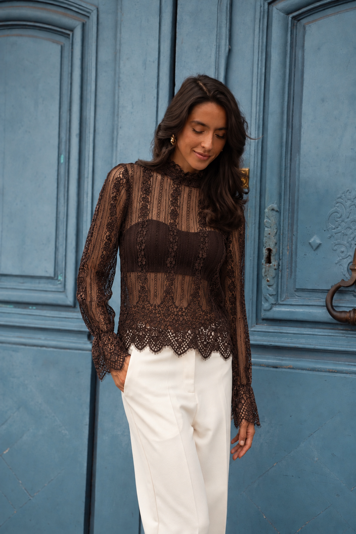 A woman wearing a sheer brown lace long-sleeve top with a structured dark brown bodice and white trousers, standing in front of a blue paneled door.