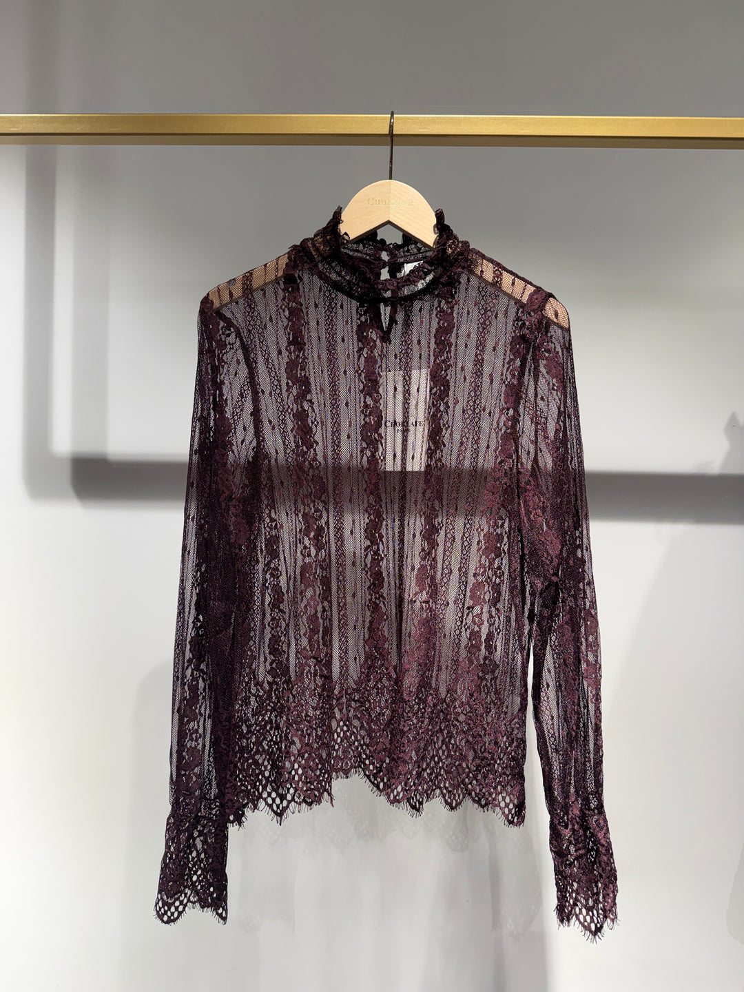 A sheer, long-sleeve lace blouse in a deep plum or burgundy shade, hanging on a wooden hanger from a gold clothing rack against a light gray wall.
