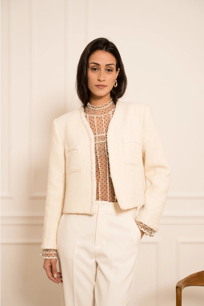 Woman wearing a cream blazer over a patterned top with white pants against a neutral background