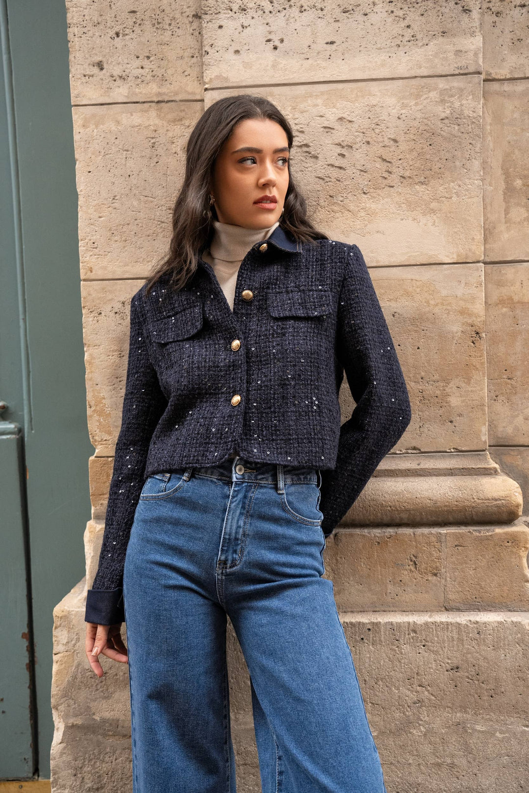 Woman wearing a dark blue textured jacket and blue jeans standing against a stone wall.