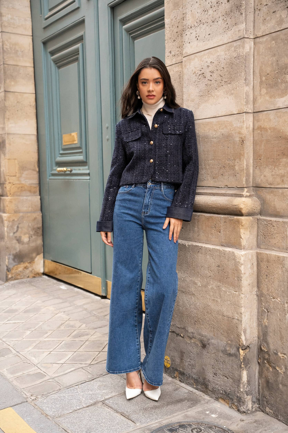 Woman in a dark denim jacket and blue jeans standing against a stone wall.