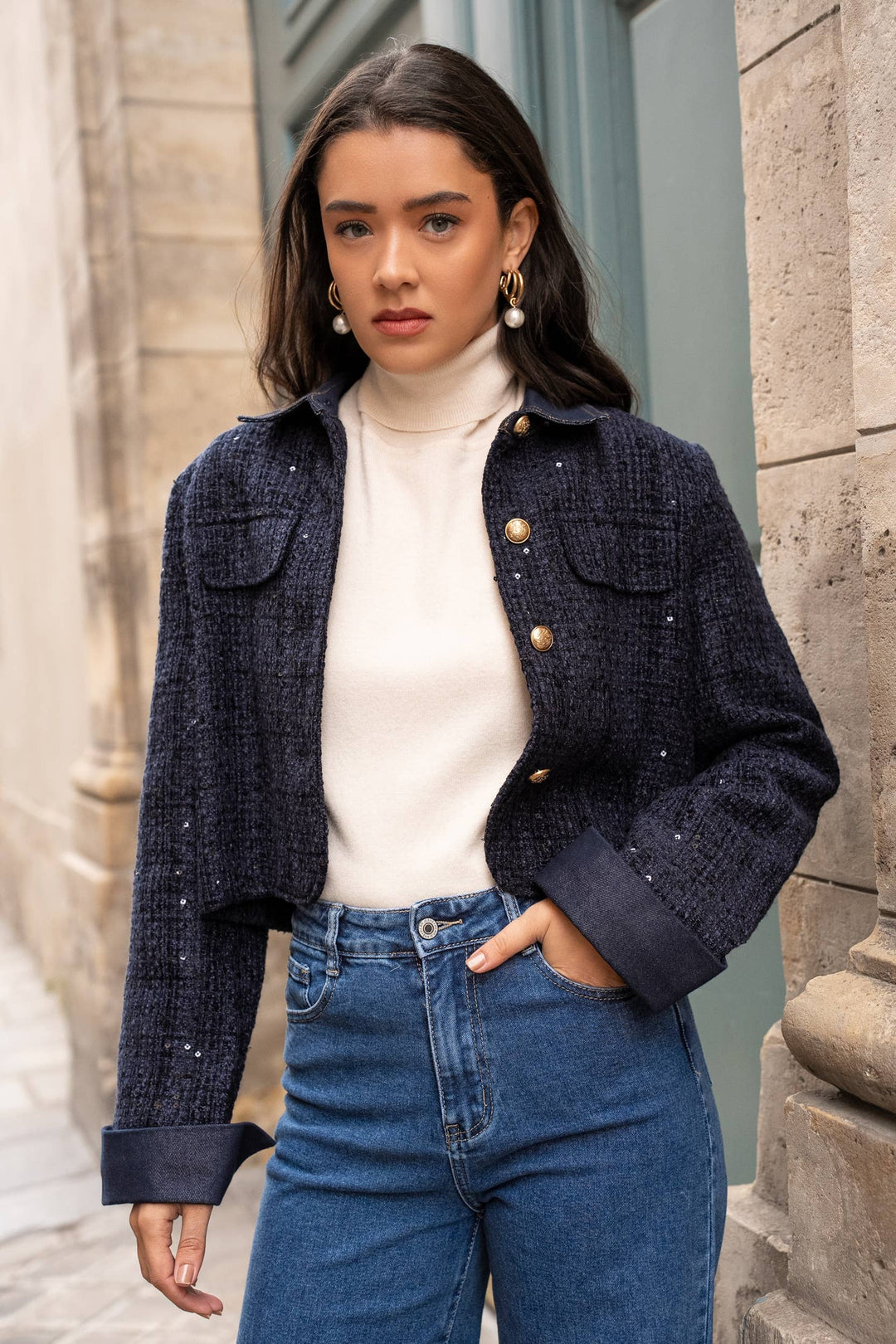 Woman wearing a dark blue textured jacket over a white turtleneck and blue jeans, standing against a stone wall.