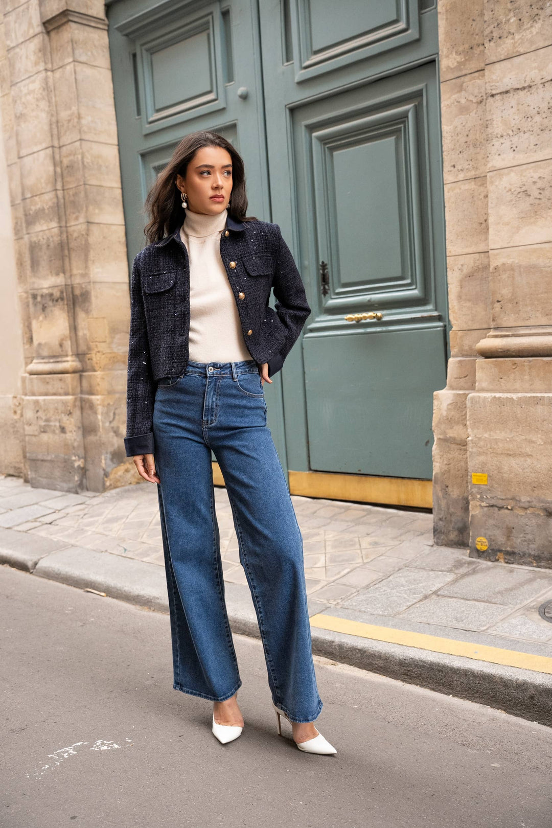 Woman in a black jacket, white turtleneck, and blue jeans standing in front of a green door.