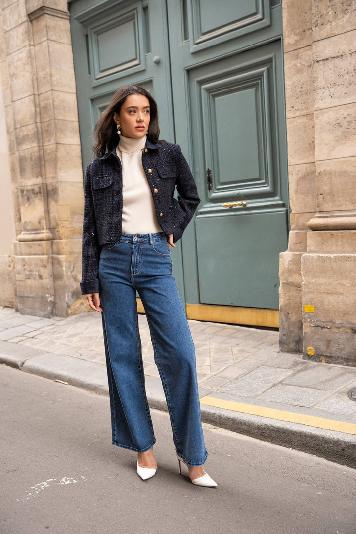 Woman in a black jacket, white turtleneck, and blue jeans standing in front of a green door.