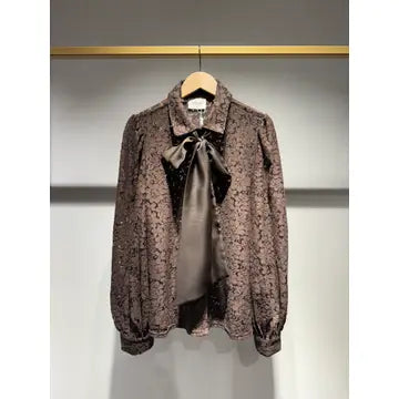 A brown patterned blouse with a large satin bow at the collar, hanging on a wooden hanger against a gray wall.