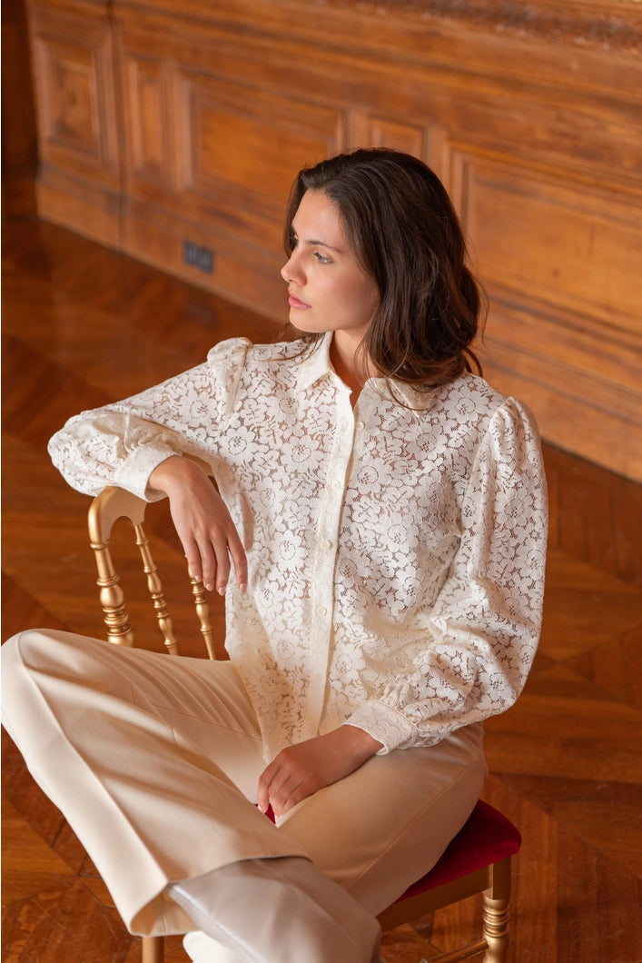 Woman wearing a white lace blouse sitting on a wooden chair in a room with wooden paneling.
