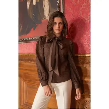 A woman wearing a dark brown long-sleeve blouse with a large bow tie neckline and white pants, standing in front of a red patterned wallpaper and wooden panel wall.