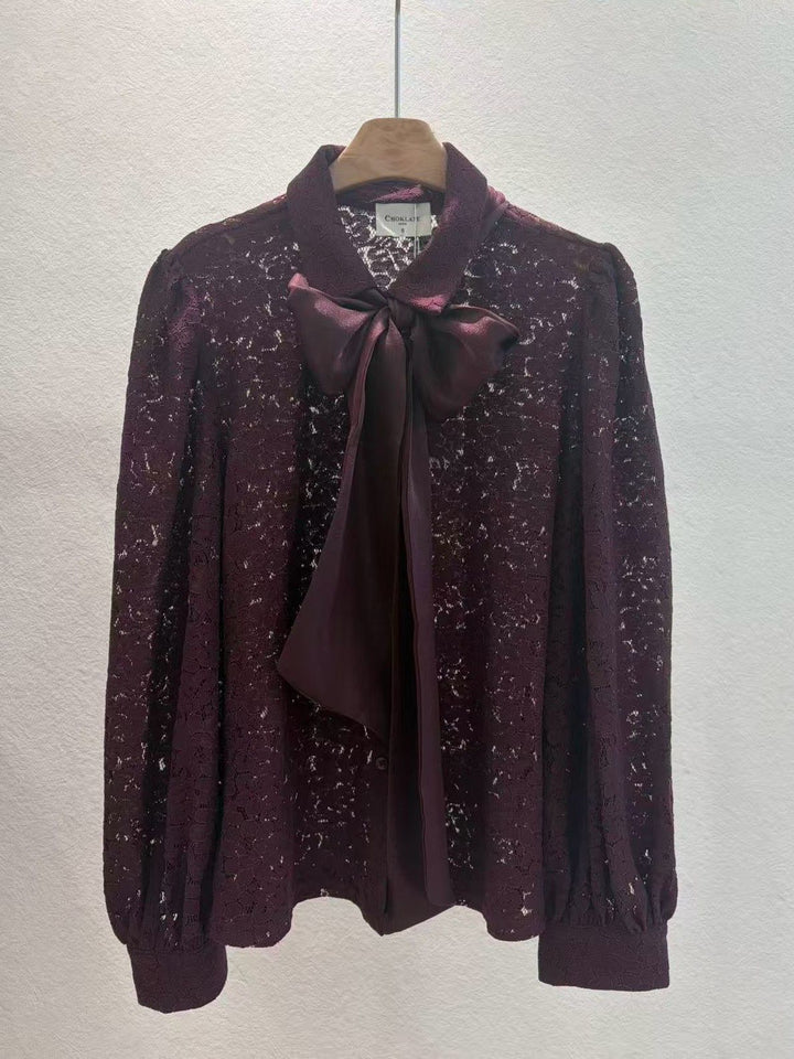 A dark plum purple long-sleeve blouse with a sheer floral lace pattern and a matching satin bow at the collar, hanging on a wooden hanger against a textured off-white wall.