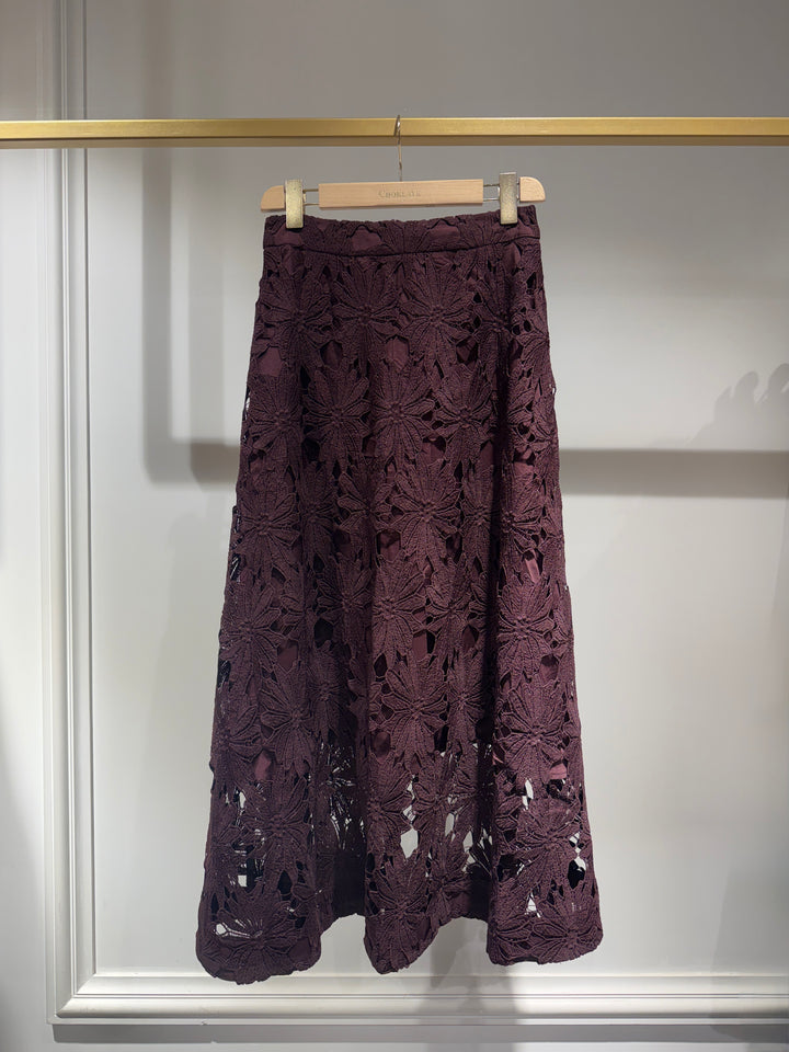 A dark plum purple lace midi skirt hanging on a wooden hanger from a gold clothing rack against a white paneled wall.
