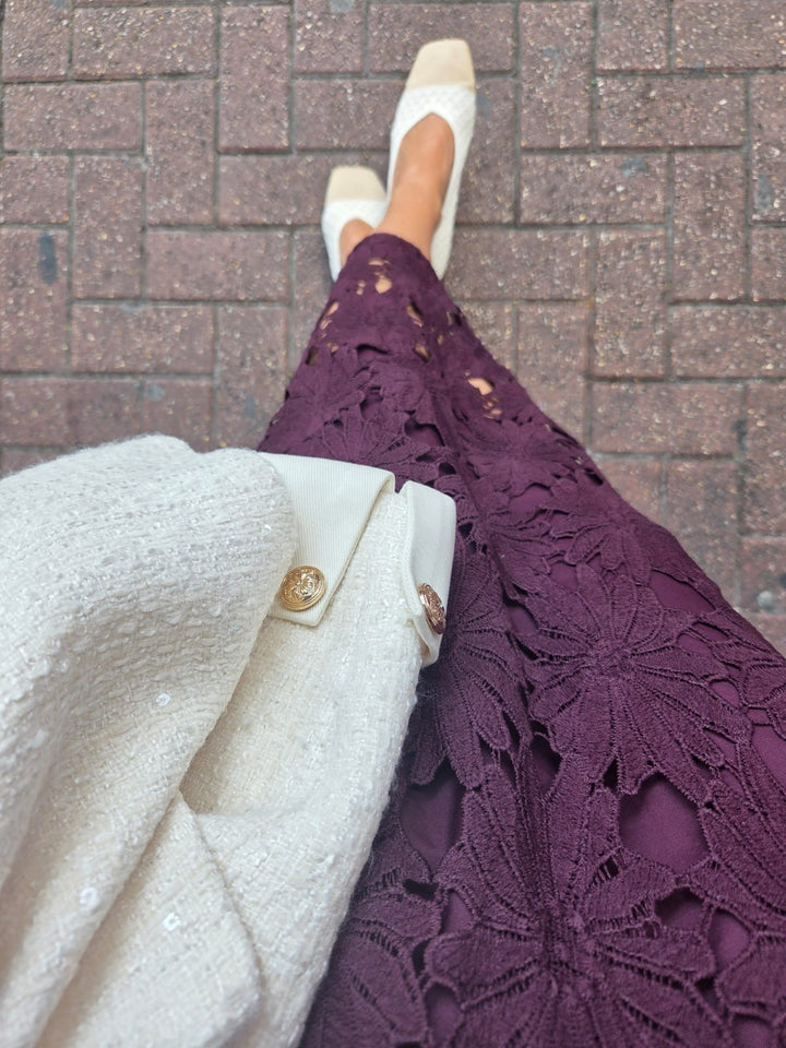 A close-up lifestyle shot of a person wearing a deep purple floral lace skirt, a cream tweed jacket with gold buttons, and cream pointed-toe flats, standing on a brick pavement.