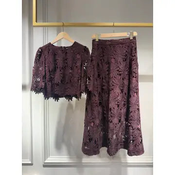 A matching two-piece women's outfit consisting of a short-sleeve lace top and a midi-length lace skirt in a deep plum purple color, hanging on wooden hangers against a white paneled wall.