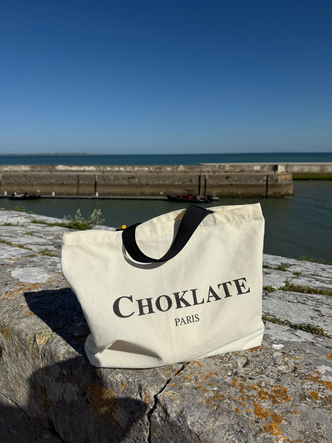 Beige tote bag with 'CHOKLATE PARIS' text on a stone ledge by a waterfront.