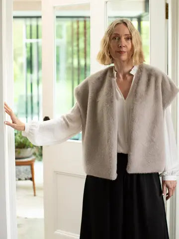 A woman wearing a light beige fuzzy sleeveless vest over a white blouse and black skirt, standing in a bright doorway with her hand resting on a doorframe.