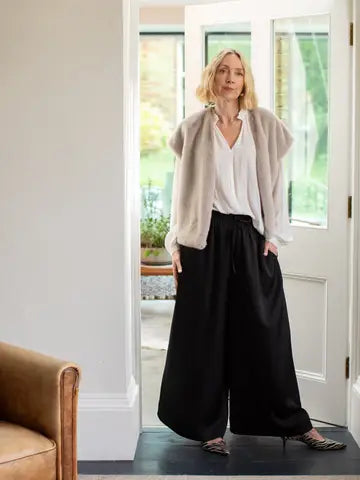 A woman stands in a doorway wearing a light beige fuzzy cardigan over a white blouse and black wide-leg pants, with patterned flats, in an indoor setting.