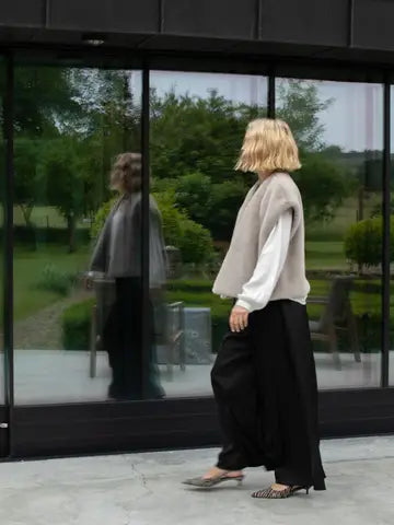 A woman with a blonde bob walks past a large glass window, wearing a beige sleeveless knit vest over a white long-sleeve blouse, black wide-leg pants, and gray slingback heels.
