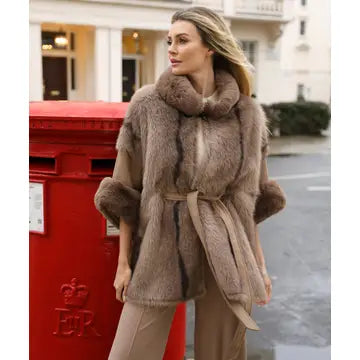 A woman wearing a brown fur wrap vest with a wide collar and tie belt, standing outdoors next to a red post box.