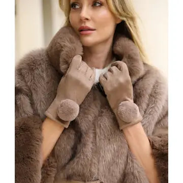 A woman wearing a plush taupe-brown faux fur coat and matching taupe-brown knit gloves with pom-pom wrist details, shown in a close-up indoor shot.