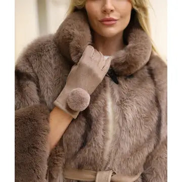 A close-up of a woman wearing a plush taupe-brown fur coat with a high collar and matching beige gloves featuring pom-pom details.