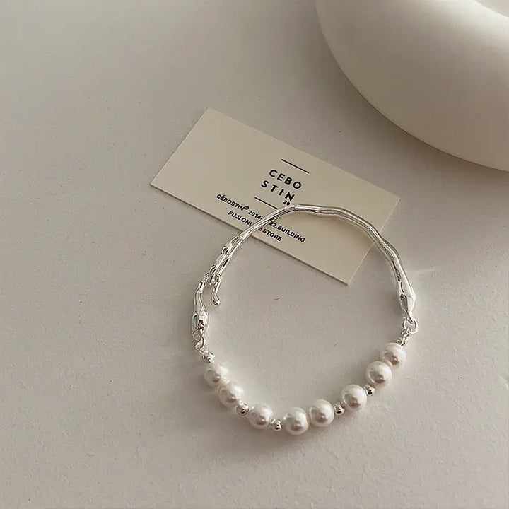 A silver-toned bracelet with a row of white faux pearls, displayed on a neutral surface next to a small card that reads 'CEBO STIN'.