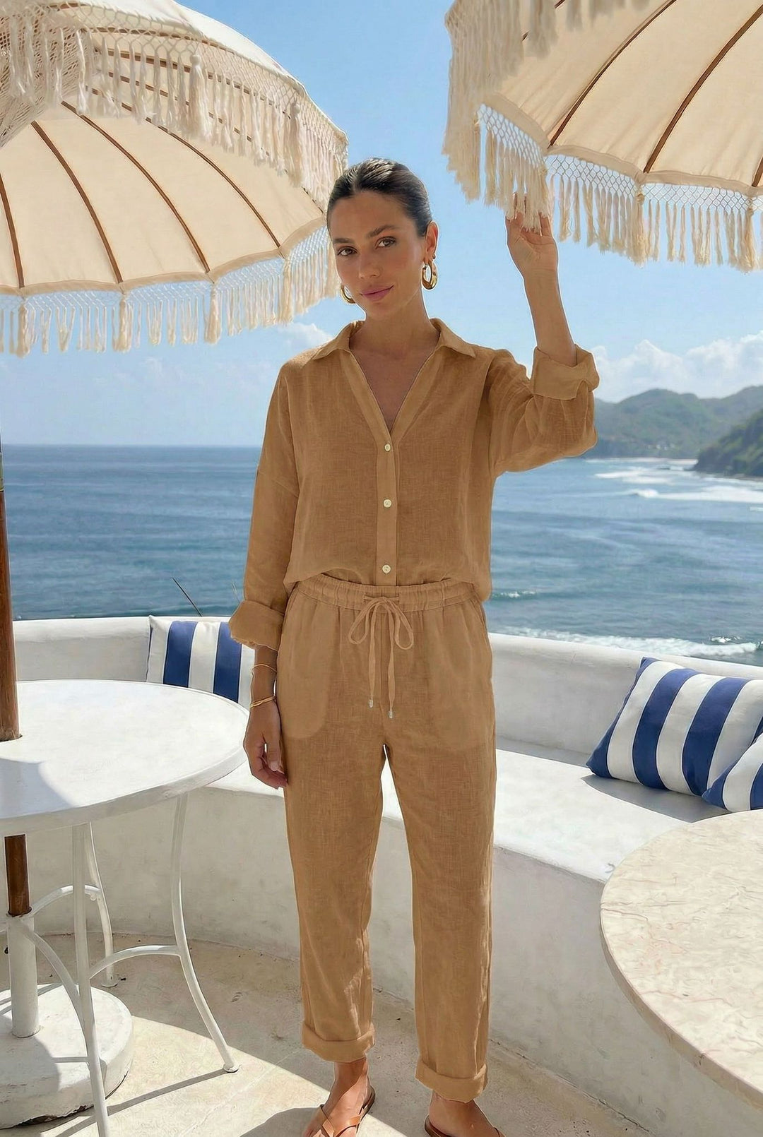 Woman in a beige outfit standing by a table with striped cushions, overlooking a scenic ocean view.
