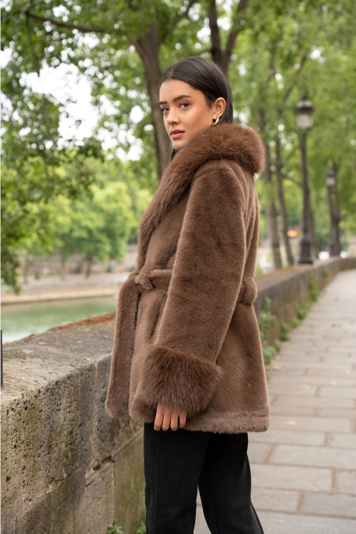Woman wearing a brown fur coat standing on a sidewalk with trees and a canal in the background