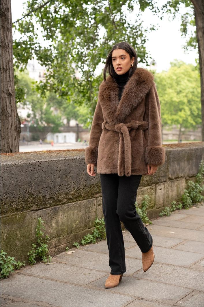 Woman wearing a brown fur coat standing on a sidewalk with greenery in the background