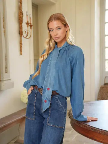 A woman wearing a blue long-sleeve blouse with a gathered neck and red button accents, paired with blue jeans, standing indoors near a wooden table.