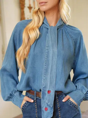 A woman wearing a blue long-sleeve blouse with a tie-neck detail and red heart-shaped buttons, paired with jeans and a brown belt.