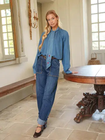A woman stands in a bright indoor room wearing a light blue denim blouse with a high neck and button front, paired with medium-wash blue jeans and dark brown shoes. A wooden table and stone floor are visible in the background.