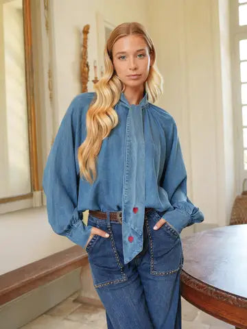 A woman modeling a blue denim long-sleeve blouse with a high gathered collar and front buttons, paired with matching blue denim jeans in an indoor room setting.
