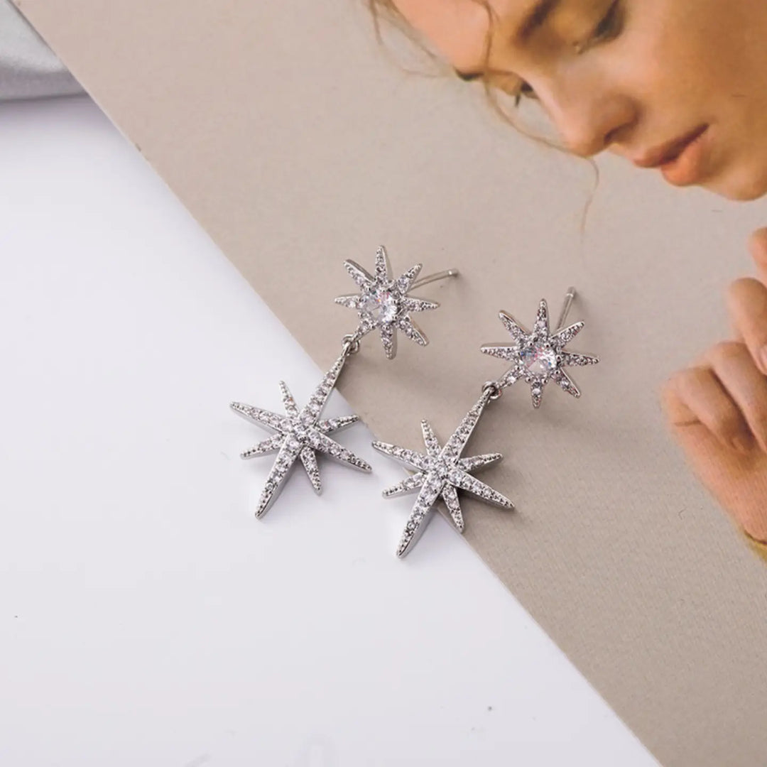 A pair of silver-toned starburst drop earrings with clear rhinestones, displayed on a split white and beige background with a model's face partially visible in the upper right.