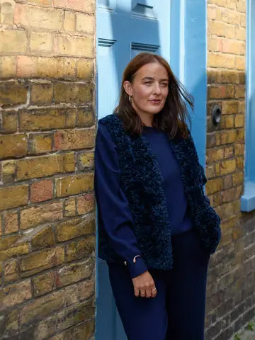 A woman with auburn hair stands leaning against a blue door framed by a yellow brick wall, wearing a navy blue long-sleeve top and matching trousers with a dark textured fuzzy vest.