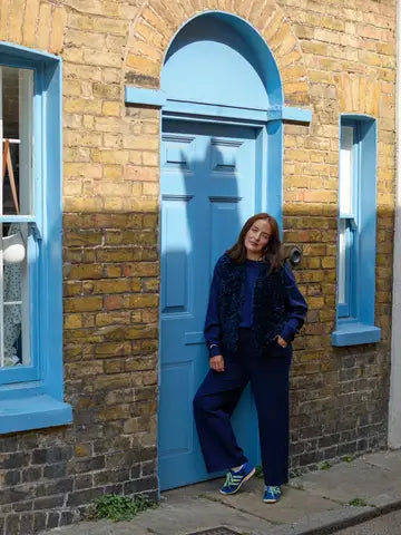 A woman stands leaning against a bright blue door set in a brick building. She is wearing a dark navy outfit with a textured dark blue jacket and blue sneakers.