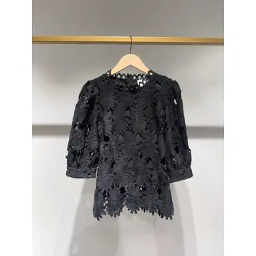 A black lace top with three-quarter sleeves and a scalloped hem, hanging on a wooden hanger against a light gray wall.
