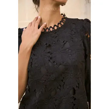 A close-up of a woman wearing a black lace top with floral embroidery and a scalloped eyelet neckline. A hand rests near the collar, and part of her neck and chin are visible.