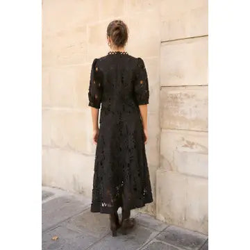 A woman viewed from behind wearing a black lace midi dress with elbow-length sleeves, standing against a beige stone wall.