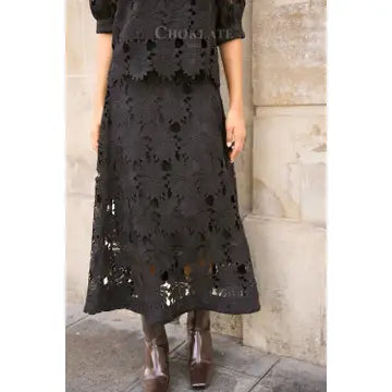 A model wearing a black lace midi skirt with a matching top, paired with brown leather boots, standing against a beige stone wall.