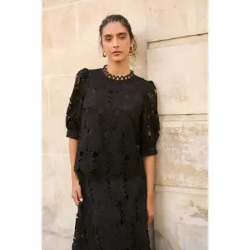 A woman wearing a black lace dress with sheer puff sleeves and a beaded neckline, standing against a light beige stone wall.