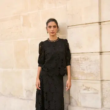 A woman wearing a black lace dress with sheer puff sleeves and a high neckline stands against a light beige stone wall.
