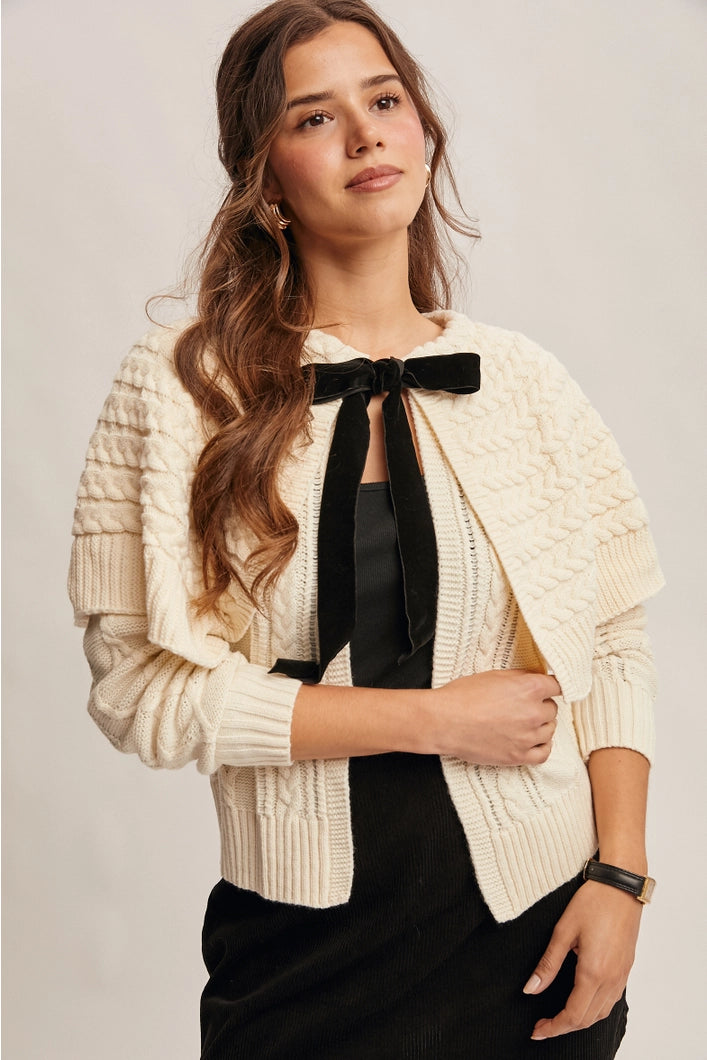 Woman wearing a cream cable knit cardigan with a black bow tie over a black top on a light gray background