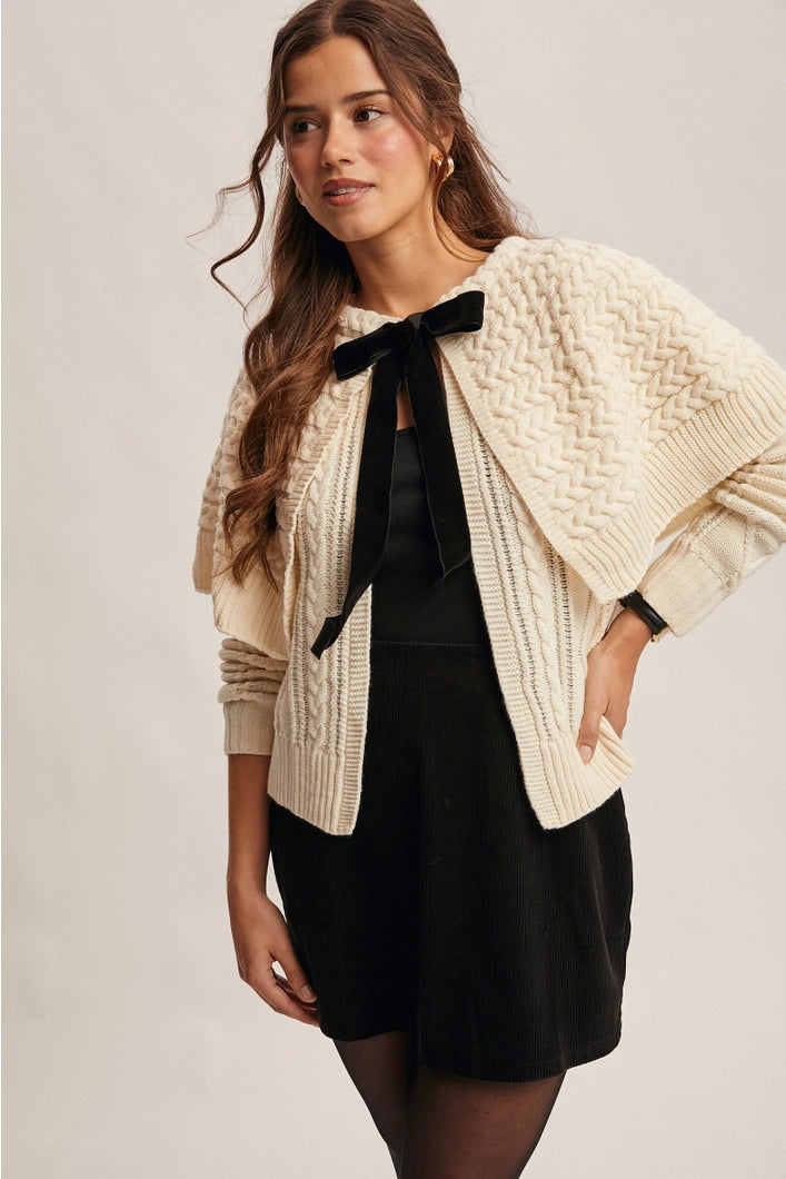 Woman wearing a beige cable knit cardigan over a black dress with a black bow tie.