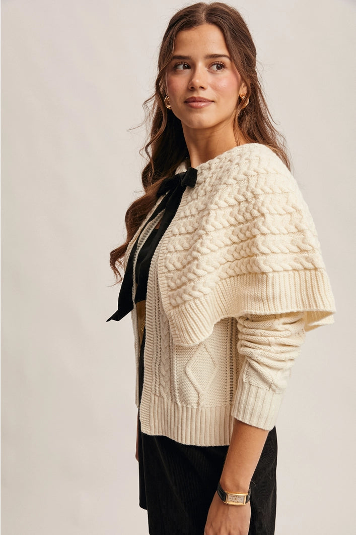 Woman wearing a cream knitted poncho with a black bow against a plain background