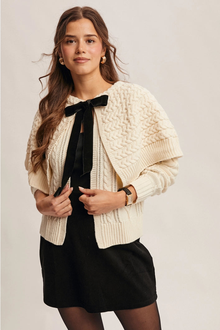 Woman wearing a cream knitted cardigan over a black dress with a plain background