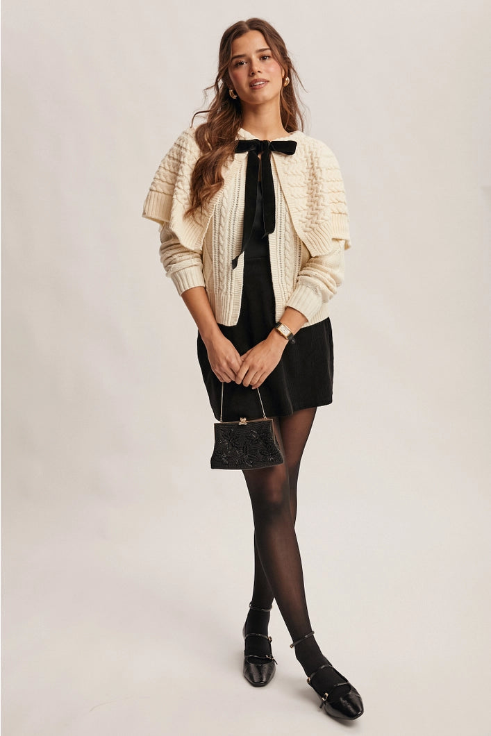 Woman wearing a cream cardigan, black dress, and black shoes on a plain background